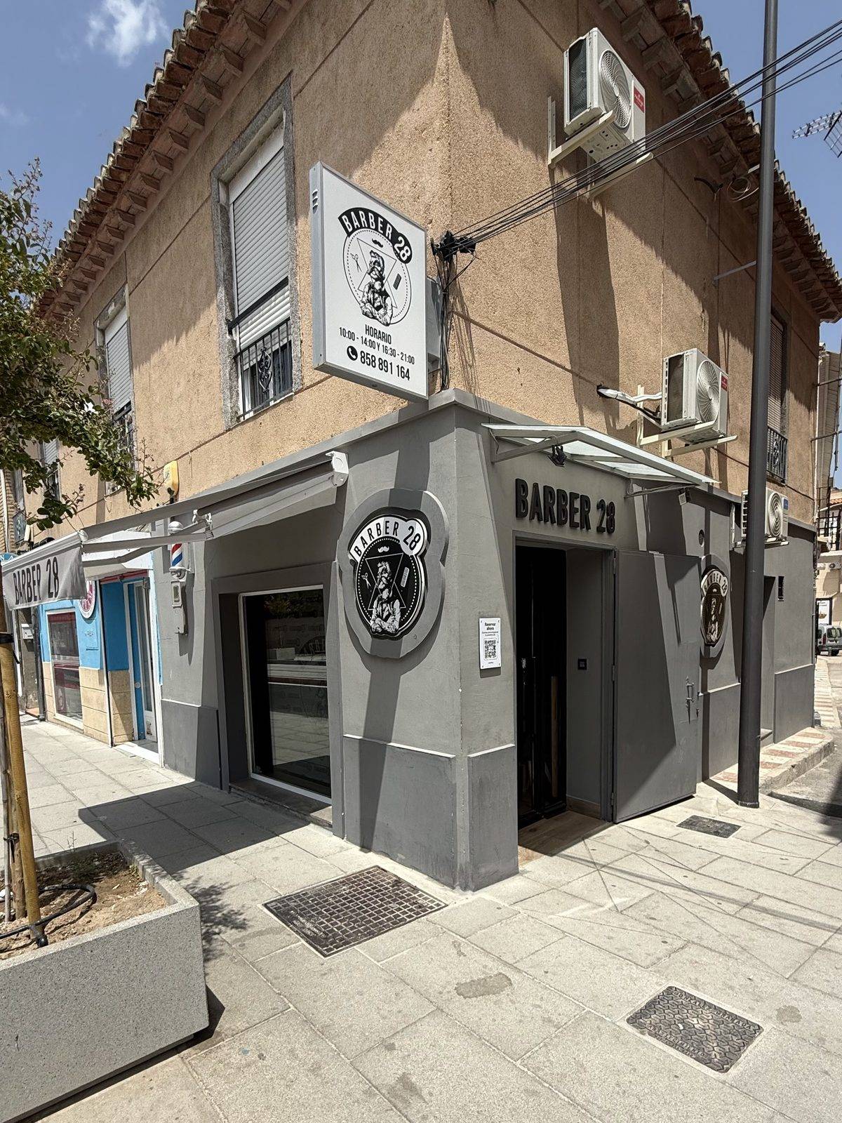 Fachada del local de Churriana de la Vega: esquina gris con el emblema de la casa, el rótulo BARBER 28 y el cartel de horario colgado en alto