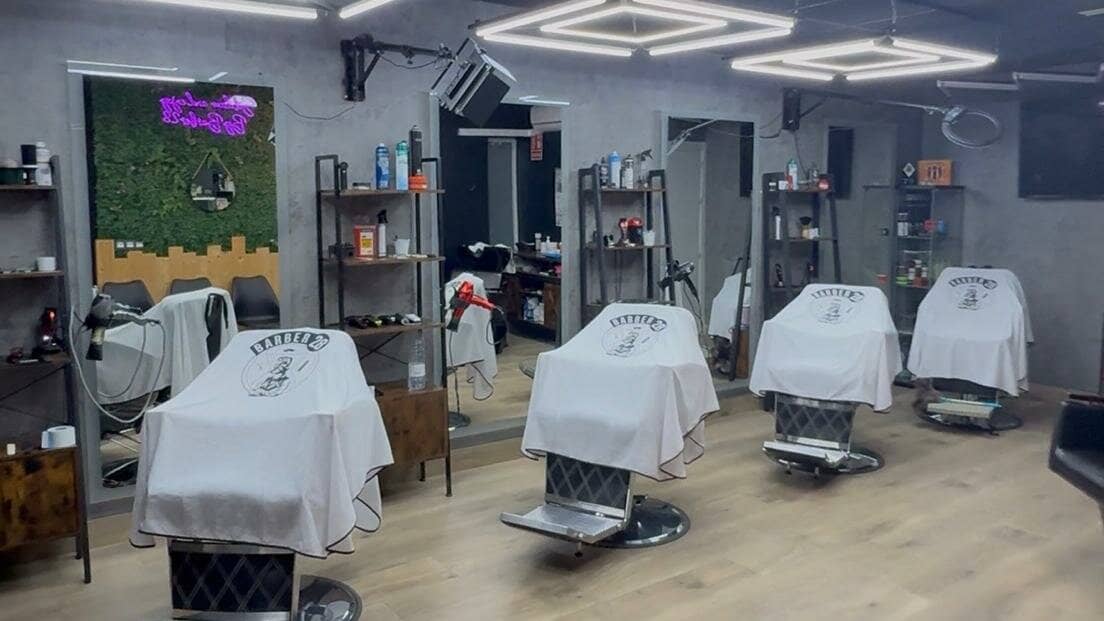 Interior del local de Churriana de la Vega: cuatro sillones de barbero cubiertos con capas blancas de Barber28 frente a los espejos, bajo lámparas geométricas